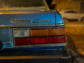 Close-up view of the rear section of a blue vintage Toyota Corona car, showing its badge and taillights. In the background, another car is slightly visible, parked on a dimly lit street.