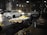 A street vendor at night organizes fruits and vegetables under a bright lamp. The scene includes a variety of produce like bananas, grapes, and oranges displayed in baskets on a cart. A blurred motion of a white car passes by on the road, while a truck is parked nearby on the dark street.