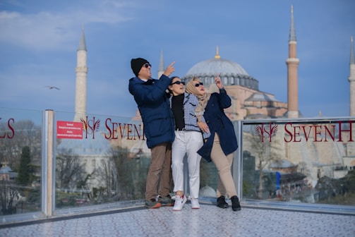 Four people stand on a platform with glass railings, with two large minarets and a dome structure visible in the background. They are casually dressed, wearing jackets and sunglasses, and appear to be enjoying a tourist spot. The sky is clear with a few clouds.