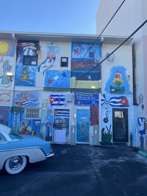 The wall is covered with vibrant murals depicting Cuban flags, tropical scenes, figures playing music, and a mix of artistic symbols and text. A classic blue and white car is parked in front of the colorful building.