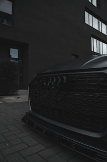 A close-up of a car's front grille with a sleek, modern design, including a distinctive logo. The setting is urban, with a brick exterior of a building and large windows in the background. The overall ambiance is moody with low lighting.