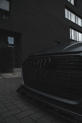 A close-up of a car's front grille with a sleek, modern design, including a distinctive logo. The setting is urban, with a brick exterior of a building and large windows in the background. The overall ambiance is moody with low lighting.