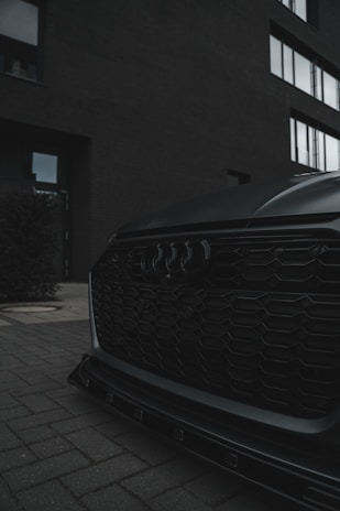A close-up of a car's front grille with a sleek, modern design, including a distinctive logo. The setting is urban, with a brick exterior of a building and large windows in the background. The overall ambiance is moody with low lighting.