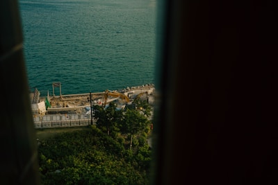 A construction site is visible near the edge of a large body of water. The scene includes construction machinery, trees, and barriers surrounding the area. The view is framed on both sides, possibly by a window or a structure.