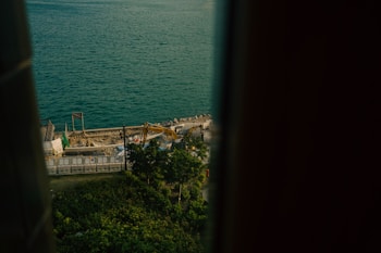 A construction site is visible near the edge of a large body of water. The scene includes construction machinery, trees, and barriers surrounding the area. The view is framed on both sides, possibly by a window or a structure.