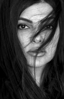 A close-up, black and white portrait of a person with long hair slightly covering the face. The expression is intense, with deep, captivating eyes. The lighting highlights the texture of the skin and the strands of hair. The composition emphasizes the elegance and mystery of the subject.