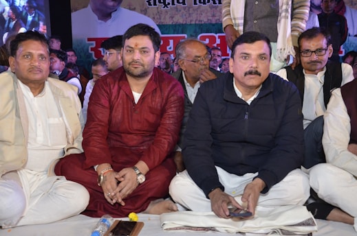 A group of people sit closely together, most of them dressed in traditional attire. The central figure in a red kurta appears focused, while others around him wear suits and casual clothing. The backdrop features a large poster with partially visible text and images.