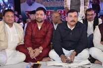 A group of people sit closely together, most of them dressed in traditional attire. The central figure in a red kurta appears focused, while others around him wear suits and casual clothing. The backdrop features a large poster with partially visible text and images.