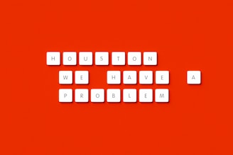 White square tiles with black letters spell out 'HOUSTON WE HAVE A PROBLEM' against a bright red background.