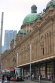 A historic building with a large green dome and intricate architectural details stands prominently. In front, a modern red and white tram travels along the street, with several people walking on the sidewalk. The juxtaposition of old and new elements creates a dynamic urban scene.