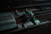 A close-up view of industrial metal machinery, featuring gears, rods, and grated surfaces. The composition includes worn and rusty elements, indicating usage and age.