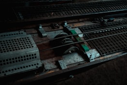 A close-up view of industrial metal machinery, featuring gears, rods, and grated surfaces. The composition includes worn and rusty elements, indicating usage and age.