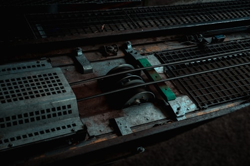 A close-up view of industrial metal machinery, featuring gears, rods, and grated surfaces. The composition includes worn and rusty elements, indicating usage and age.