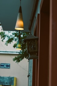 A dimly lit hanging lantern and two pendant lights in an outdoor setting. The lantern is attached to a red wall, and there is a soft glow from the lights. A tree and a building facade are visible in the background, creating a calm and serene atmosphere.