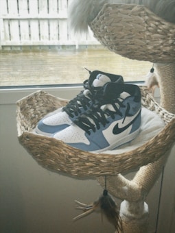A pair of sneakers with a white and gray color scheme sits on a woven basket-like platform. The sneakers have black laces and a recognizable swoosh on the side. The basket is part of a cat tree structure, situated in front of a window. The setting suggests an indoor environment with natural light coming through.