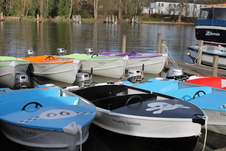 Colorful rowboats with distinctive names and designs are moored at a wooden dock on a calm lake, surrounded by trees and a distant view of a house. The boats are equipped with small outboard motors.