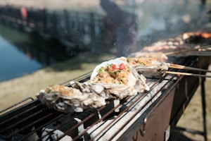 Several oysters filled with a colorful mixture of cooked ingredients are placed on skewers over a grill. The scene takes place outdoors near a body of water. Steam rises from the food, indicating they are being grilled or smoked. The background is slightly blurred, focusing attention on the oysters.