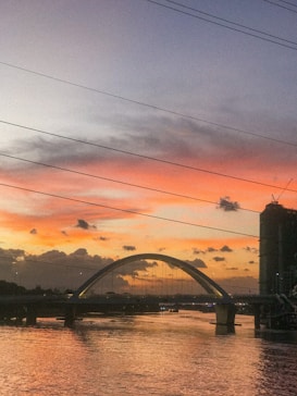 A scenic view of a river at dusk, with a prominent arch bridge crossing over. The sky displays hues of orange and pink, creating a beautiful sunset. There are silhouetted clouds scattered across the horizon, and a tall, partially constructed building is visible on the right. Electrical wires stretch across the sky, adding an urban element.