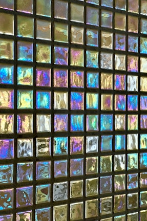 A surface composed of small, square, reflective tiles displaying an iridescent effect, creating a colorful and shimmering appearance. The squares are arrayed in a grid pattern, and the light reflects off each tile, producing a spectrum of colors including blues, purples, yellows, and greens.