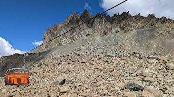 A rugged mountainous landscape featuring rocky terrain and a sharp, jagged ridge under a clear blue sky. A cable car with a protective orange cover is suspended by a cable.