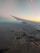 Aerial view from an airplane, showing the wing extending over a landscape dotted with organized agricultural fields. The horizon is visible with a gradient sky, transitioning from soft blues to warm yellows and oranges, indicating a sunrise or sunset.