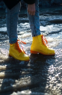 A pair of bright yellow boots with orange laces is worn by a person standing on a wet, icy surface. The person is wearing light blue jeans, and the surrounding area reflects the sun, creating a sparkling effect on the ice.