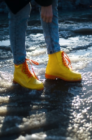 A pair of bright yellow boots with orange laces is worn by a person standing on a wet, icy surface. The person is wearing light blue jeans, and the surrounding area reflects the sun, creating a sparkling effect on the ice.
