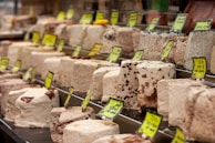 A display of various blocks of halva, featuring different colors and textures, with yellow price tags. The halva blocks are arranged neatly in rows, showcasing different types and flavors.