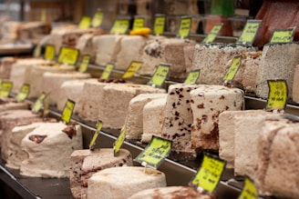 A display of various blocks of halva, featuring different colors and textures, with yellow price tags. The halva blocks are arranged neatly in rows, showcasing different types and flavors.