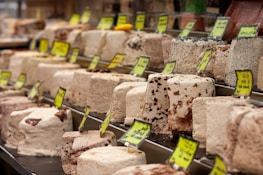 A display of various blocks of halva, featuring different colors and textures, with yellow price tags. The halva blocks are arranged neatly in rows, showcasing different types and flavors.