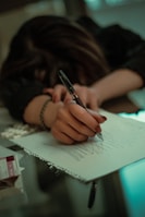 A person with long hair is resting their head on a table while holding a pen and scribbling on a notepad. The environment appears dimly lit, creating a moody atmosphere. There are some papers and a small box nearby on the table.