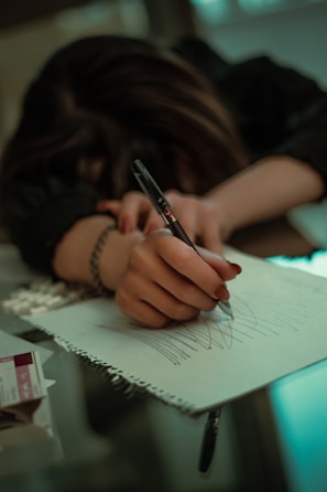 A person with long hair is resting their head on a table while holding a pen and scribbling on a notepad. The environment appears dimly lit, creating a moody atmosphere. There are some papers and a small box nearby on the table.
