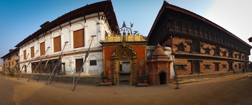 Traditional architectural buildings featuring ornate carvings and wooden details, with support beams visible along the front. The structures combine classic white and brick-red facades, and an intricately decorated entrance gate. The scene is lit by warm, natural light.