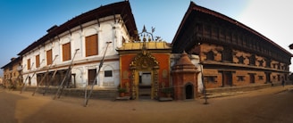 Traditional architectural buildings featuring ornate carvings and wooden details, with support beams visible along the front. The structures combine classic white and brick-red facades, and an intricately decorated entrance gate. The scene is lit by warm, natural light.
