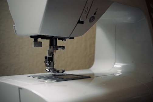 A close-up view of a sewing machine, focusing on the needle and presser foot area. The machine is mostly white, with some metallic components visible. The background is neutral, emphasizing the machine itself.