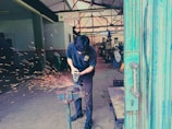 A person is using a grinding tool on a metal object clamped in a vice, creating bright orange sparks in an industrial workshop. In the background, metalworking machinery and equipment are visible, with another individual sitting in the distance. The atmosphere appears busy and functional, with a focus on craftsmanship and metalwork.