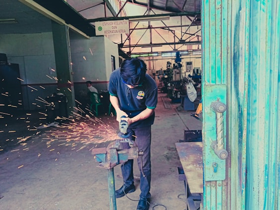 A person is using a grinding tool on a metal object clamped in a vice, creating bright orange sparks in an industrial workshop. In the background, metalworking machinery and equipment are visible, with another individual sitting in the distance. The atmosphere appears busy and functional, with a focus on craftsmanship and metalwork.