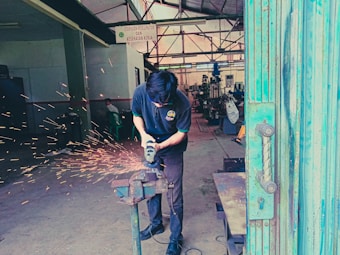 A person is using a grinding tool on a metal object clamped in a vice, creating bright orange sparks in an industrial workshop. In the background, metalworking machinery and equipment are visible, with another individual sitting in the distance. The atmosphere appears busy and functional, with a focus on craftsmanship and metalwork.