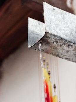 A close-up image of a metal gutter with water flowing from its edge. Below the gutter, colorful strings with beads and decorations hang, featuring shades of red, yellow, and black. The background is out of focus, creating a contrast with the detailed metal surface and the vibrant hanging ornaments.