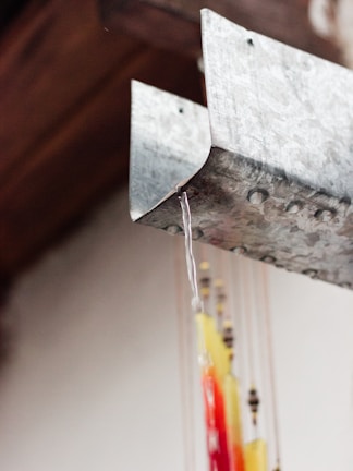 A close-up image of a metal gutter with water flowing from its edge. Below the gutter, colorful strings with beads and decorations hang, featuring shades of red, yellow, and black. The background is out of focus, creating a contrast with the detailed metal surface and the vibrant hanging ornaments.