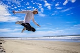 A person in athletic clothing is performing an impressive jump against a backdrop of a sandy beach and a bright blue sky scattered with clouds. The beach extends along the horizon with gentle waves meeting the shore. The person appears to be airborne, with arms spread and knees bent, creating a dynamic and energetic pose.