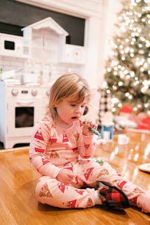 A small child in festive pajamas sits on a wooden floor, appearing to brush their teeth with a colorful toothbrush. In the background, there is a decorated Christmas tree and a play kitchen set.