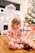 A small child in festive pajamas sits on a wooden floor, appearing to brush their teeth with a colorful toothbrush. In the background, there is a decorated Christmas tree and a play kitchen set.