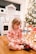 A small child in festive pajamas sits on a wooden floor, appearing to brush their teeth with a colorful toothbrush. In the background, there is a decorated Christmas tree and a play kitchen set.