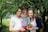 A family consisting of a man, woman, and a child standing closely together outdoors. The man is wearing glasses and a white t-shirt, while the woman is dressed in a white top with a dark sweater. The child is holding a plant and dressed in a grey top and red pants. They are surrounded by lush green trees and foliage.