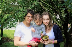 A family consisting of a man, woman, and a child standing closely together outdoors. The man is wearing glasses and a white t-shirt, while the woman is dressed in a white top with a dark sweater. The child is holding a plant and dressed in a grey top and red pants. They are surrounded by lush green trees and foliage.