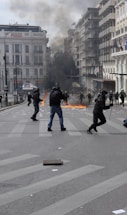 Protesters are actively engaged in a street demonstration, with some individuals running through the street and others confronting police. Flames are visible on the ground, and thick smoke rises, indicating possibly burning objects. The scene suggests a chaotic or tense environment in an urban setting, with several buildings lining the street and some flags visible on one building.