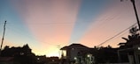 A residential area at dusk with silhouetted structures against a gradient sky. Power lines stretch across the scene, intersecting in the foreground. The sky transitions from a warm orange near the horizon to a cool blue at the top, casting serene light across the landscape.