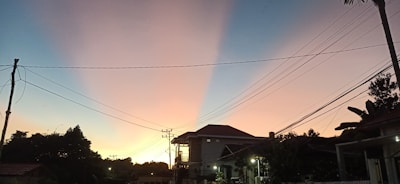 A residential area at dusk with silhouetted structures against a gradient sky. Power lines stretch across the scene, intersecting in the foreground. The sky transitions from a warm orange near the horizon to a cool blue at the top, casting serene light across the landscape.