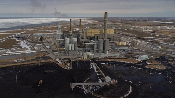 An industrial power plant with several smokestacks and large buildings is situated in a flat, expansive landscape. The surrounding area has patches of snow and water, with a network of roads and utility lines visible. An area of dark earth or coal is prominent in the foreground.